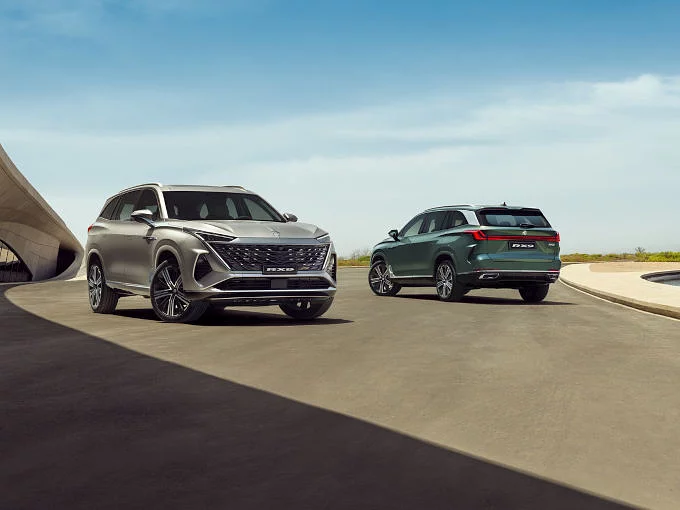 Two MG RX9 7-seater SUVs — one silver, one green — driving side by side on an open road with a modern architectural backdrop, showcasing the flagship model's bold exterior design.